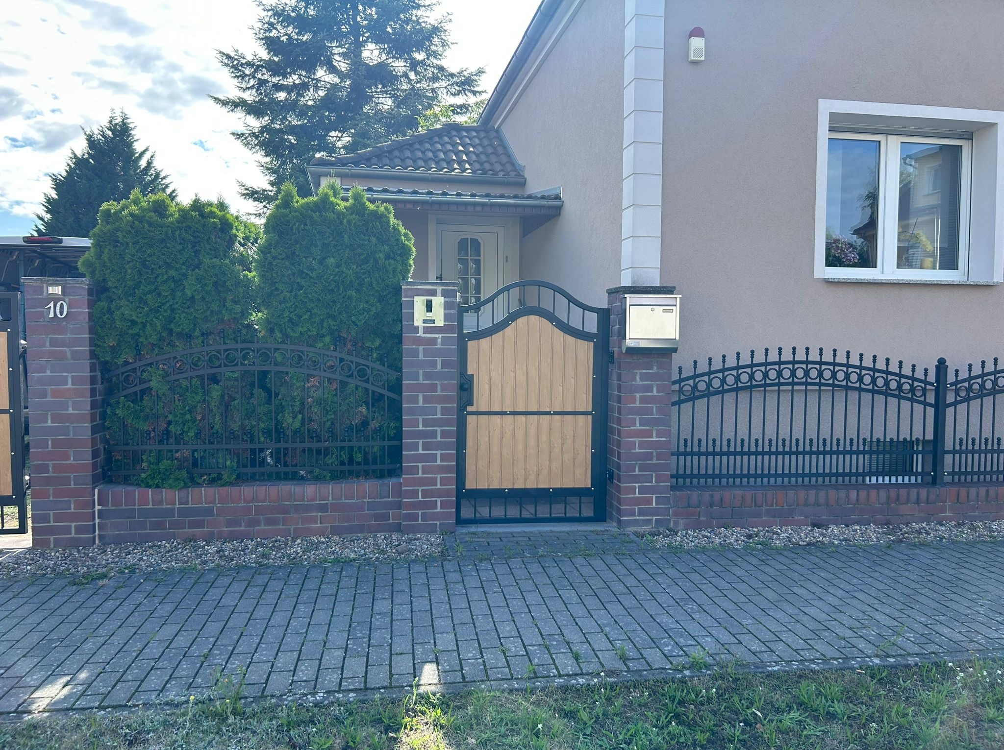 Brama z drewnianym wypełnieniem i czarnym, kutym obramowaniem, wkomponowana w ceglany murek z metalowym ogrodzeniem. Widok na dom z oknem i roślinami.