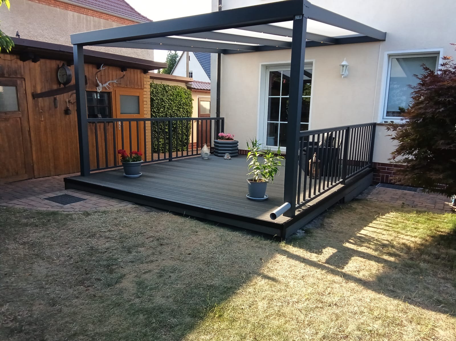 Nowoczesny taras kompozytowy z czarną pergolą i balustradą, otoczony zielenią. Widoczne donice z kwiatami i elementy dekoracyjne, tworzące przytulną przestrzeń.