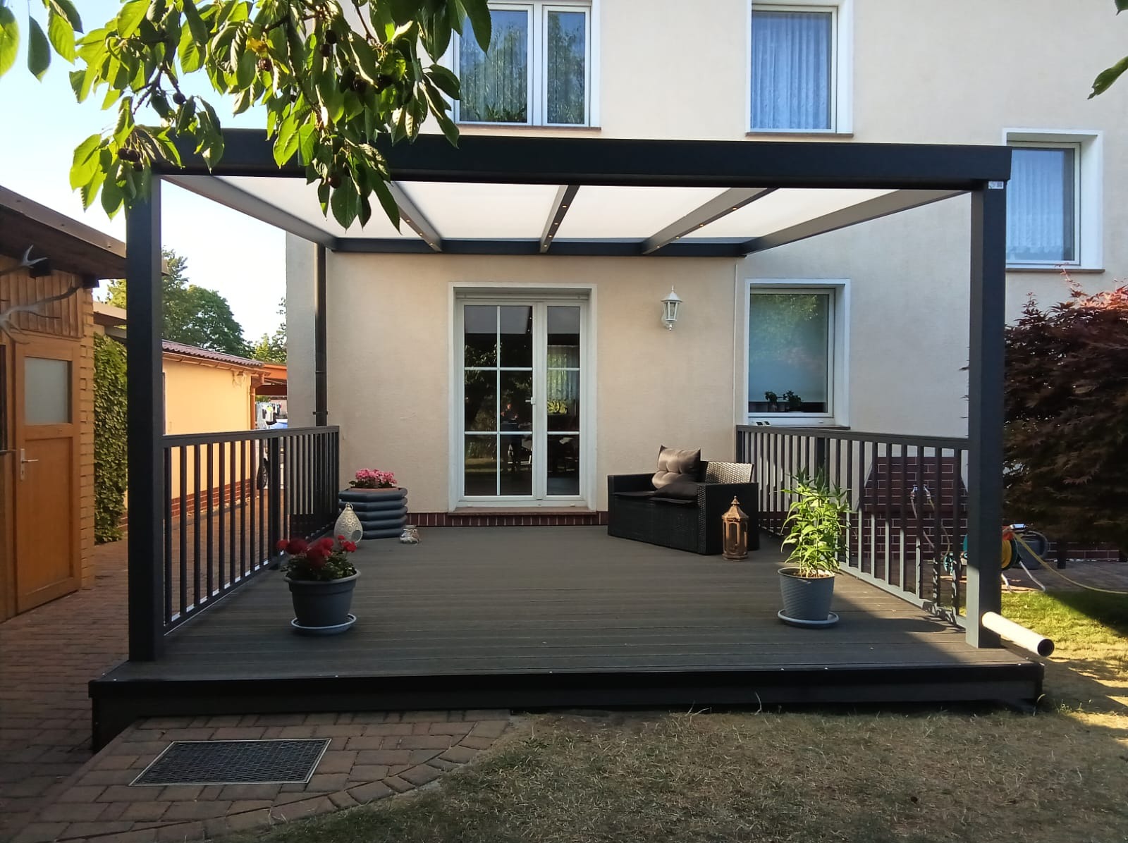 Nowoczesny taras kompozytowy z czarną pergolą i balustradą. Ciemna podłoga tarasu, meble ogrodowe, donice z kwiatami. Jasna elewacja domu w tle.