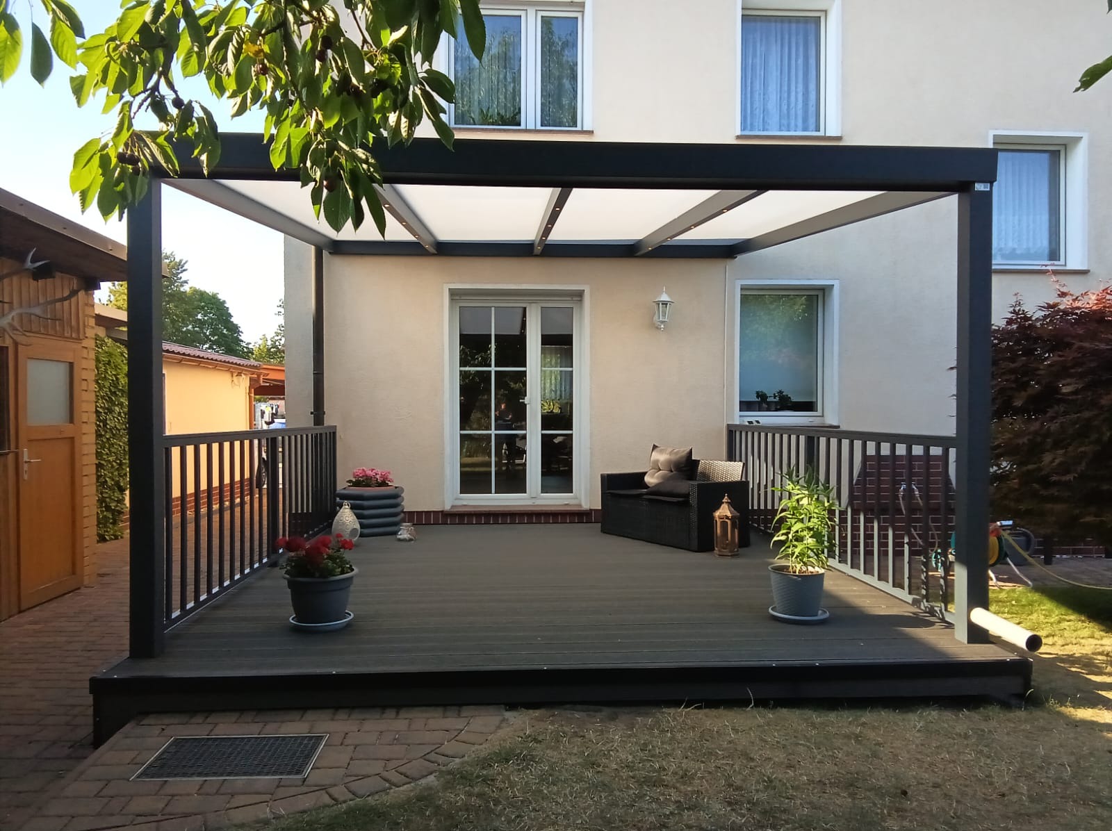 Nowoczesny taras z pergolą przy domu. Ciemna deska kompozytowa, minimalistyczne meble ogrodowe, donice z kwiatami. Oświetlenie punktowe w pergoli.