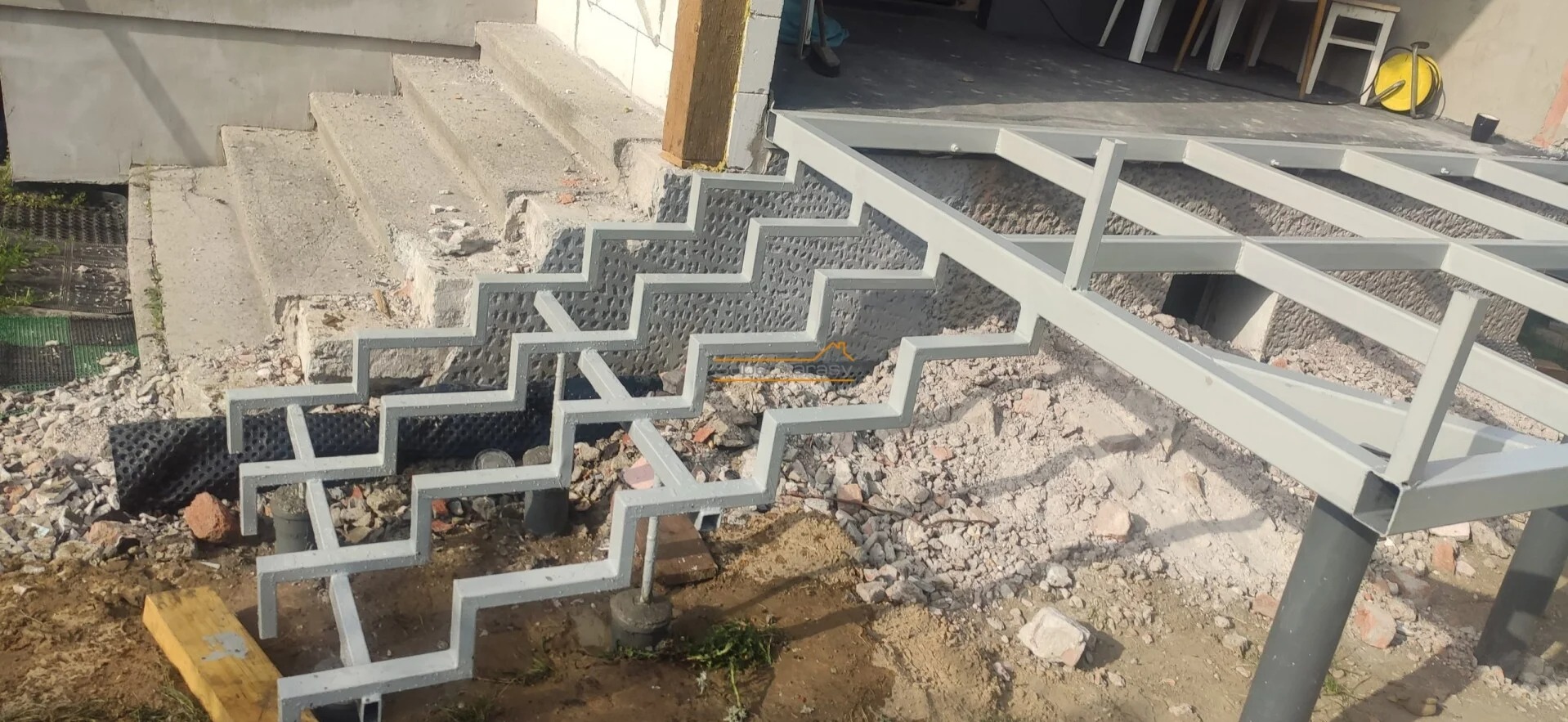 Stalowa konstrukcja schodów i tarasu w trakcie montażu. Surowe betonowe schody w tle. Widoczne elementy mocujące. Prace w toku, teren przygotowany pod budowę.