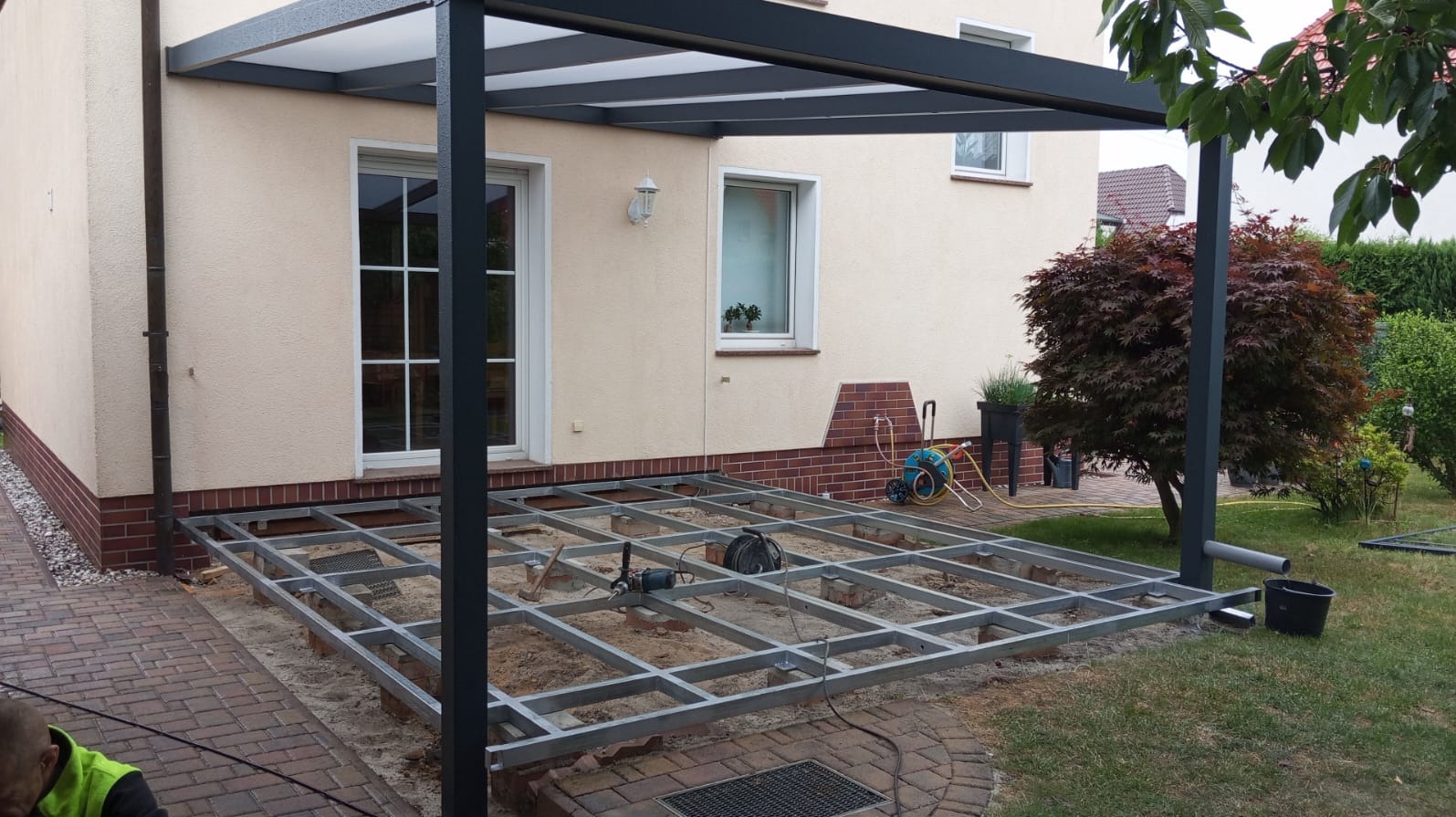 Metalowa konstrukcja tarasu kompozytowego w trakcie montażu, widoczna na tle domu z oknami i zieleni. Etap budowy z narzędziami.