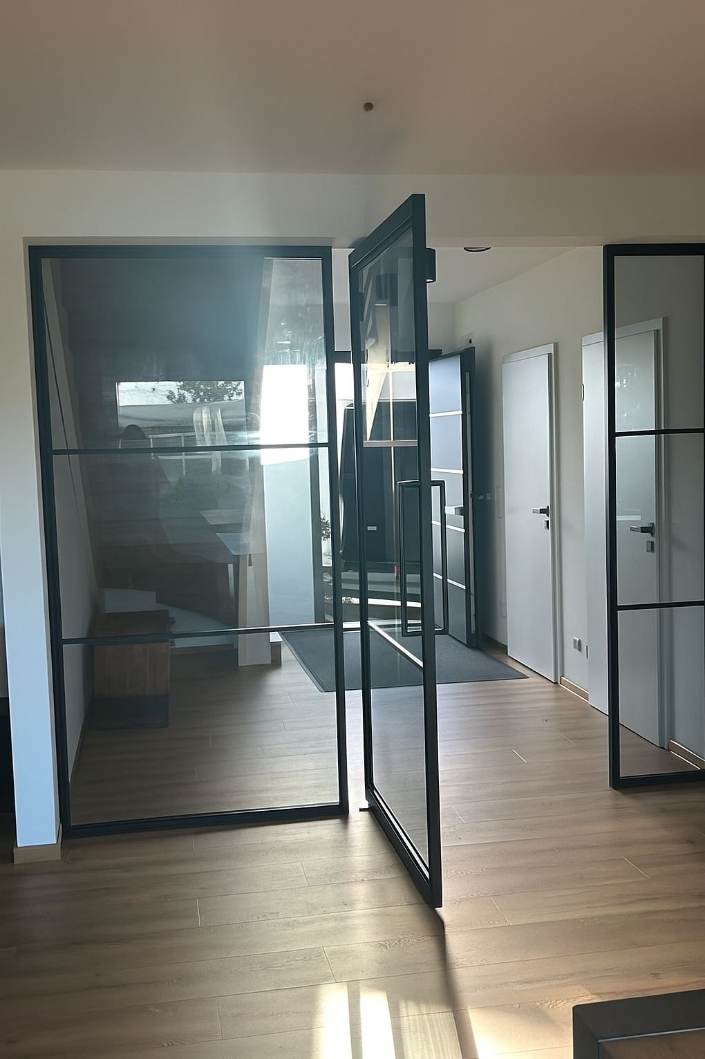 Nowoczesne, przeszklone drzwi loftowe z czarną ramą, częściowo otwarte, dzielące przestrzeń w jasnym wnętrzu z parkietem. Widoczny fragment schodów i dalsze pomieszczenia.