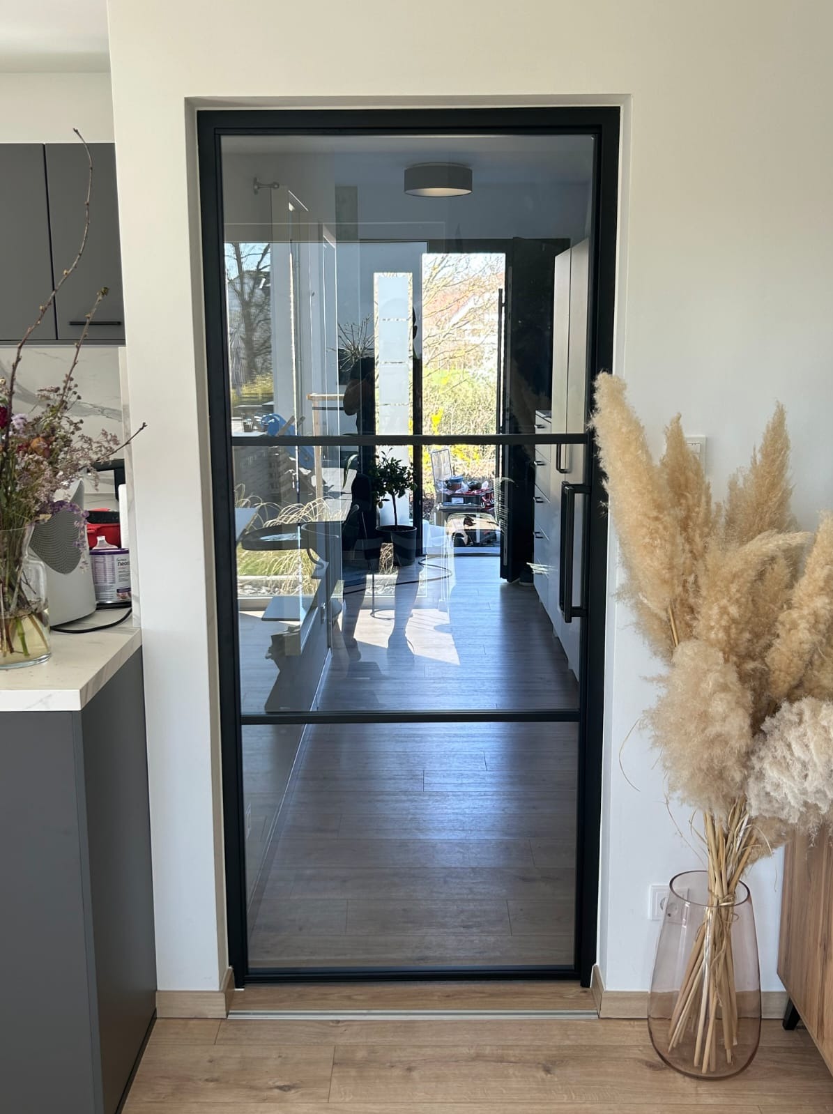 Szklane drzwi z czarną ramą, widok z kuchni na jasny korytarz. Minimalistyczny design, drewniana podłoga, dekoracje z suszonych traw. Nowoczesne wnętrze.
