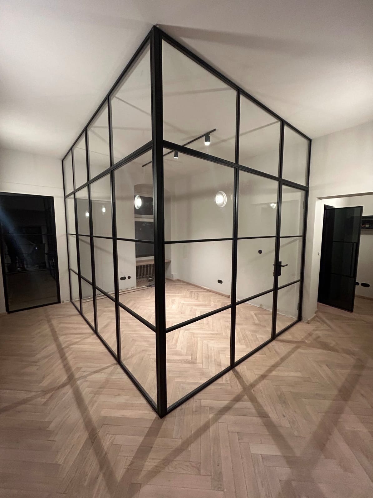 Nowoczesna, przeszklona ścianka działowa z czarnymi profilami w jasnym wnętrzu. Parkiet w jodełkę, minimalistyczne oświetlenie. Widoczne drzwi z czarną ramą.