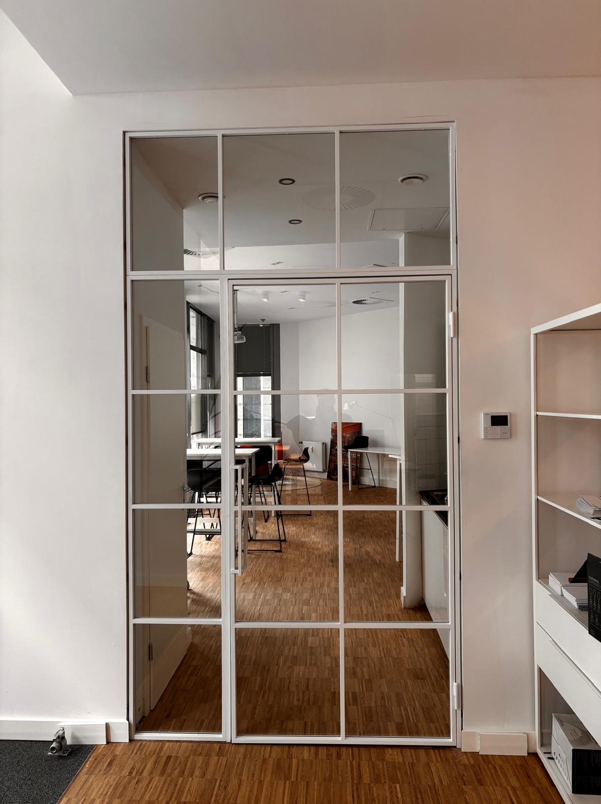 Białe, przeszklone drzwi w metalowej ramie, dzielące przestrzeń biurową z drewnianą podłogą i widocznymi meblami. Nowoczesny design, jasne wnętrze.