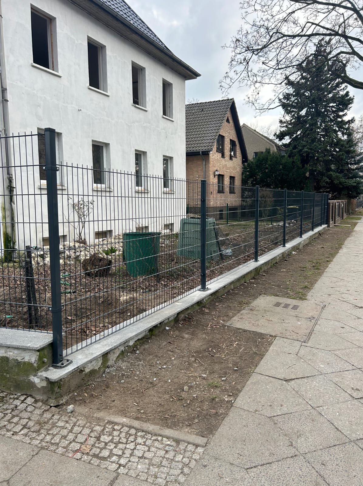 Nowoczesne ogrodzenie panelowe w kolorze grafitowym oddziela posesję od chodnika. Widoczne słupki i siatka, w tle budynki mieszkalne i drzewo.