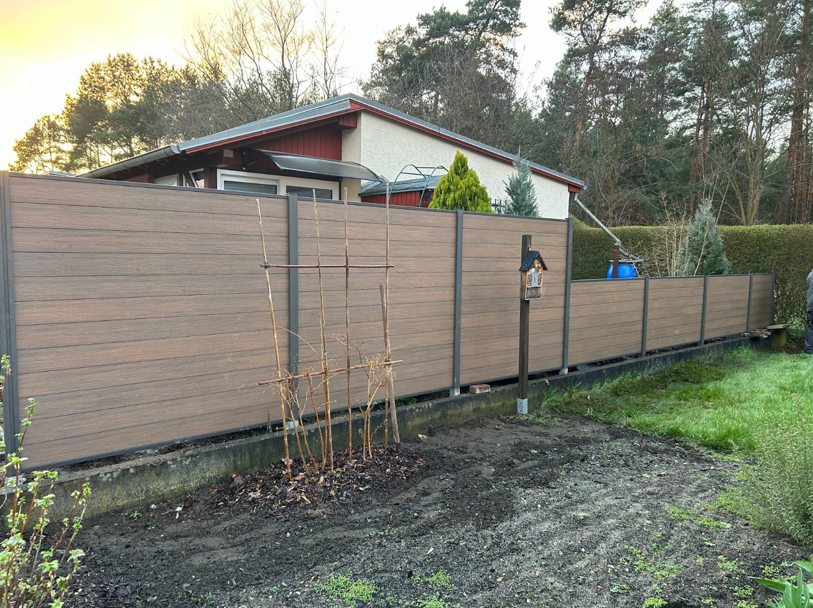 Nowoczesne ogrodzenie kompozytowe w kolorze drewna z metalowymi słupkami, widok z zewnątrz posesji, z elementami ogrodu i domu w tle.