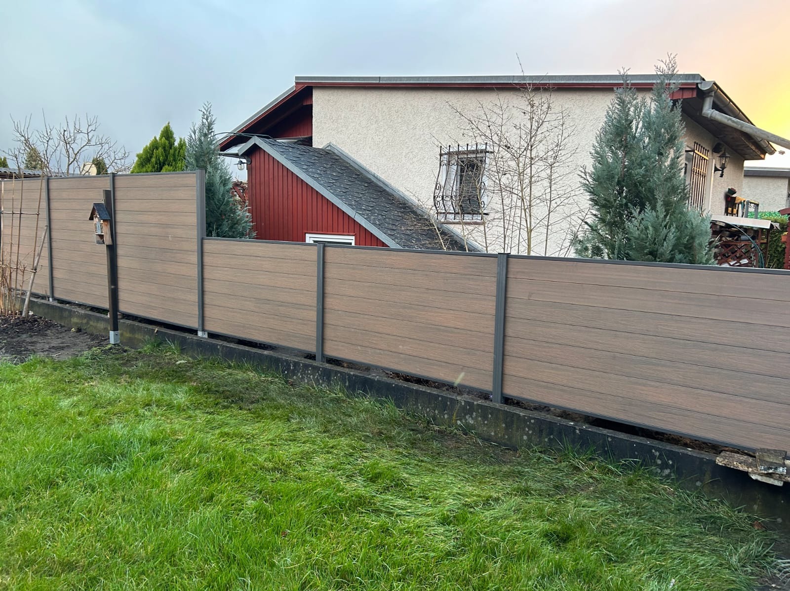 Nowoczesne ogrodzenie kompozytowe w kolorze drewna z metalowymi słupkami, widok z niskiej perspektywy na tle domu i zielonego trawnika.