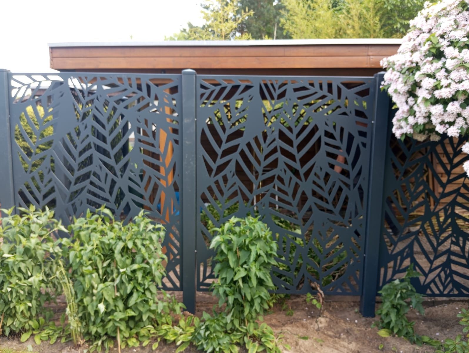 Nowoczesne ogrodzenie panelowe z motywem liści, w kolorze grafitowym, z widocznymi roślinami przed i za nim, oraz fragmentem drewnianej konstrukcji w tle.