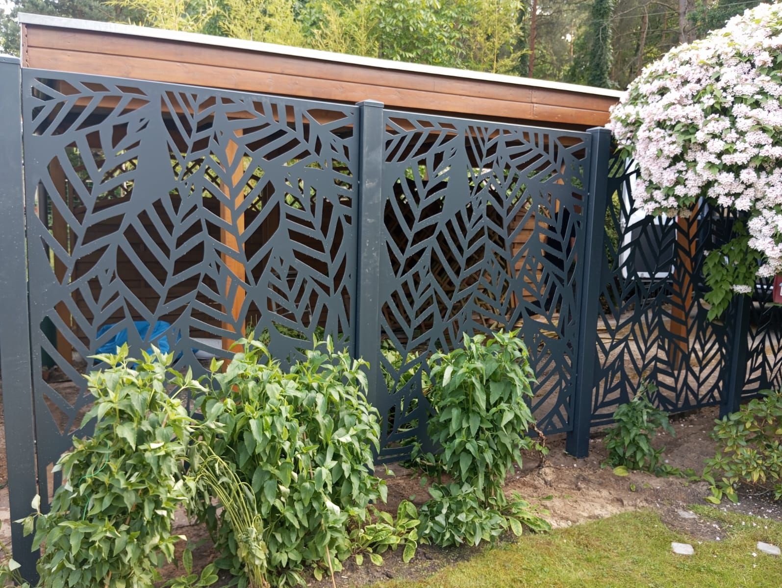 Dekoracyjne, ażurowe ogrodzenie w kolorze antracytu z motywem liści na tle drewnianej konstrukcji i zieleni. Nowoczesny design, podkreślony przez roślinność w ogrodzie.