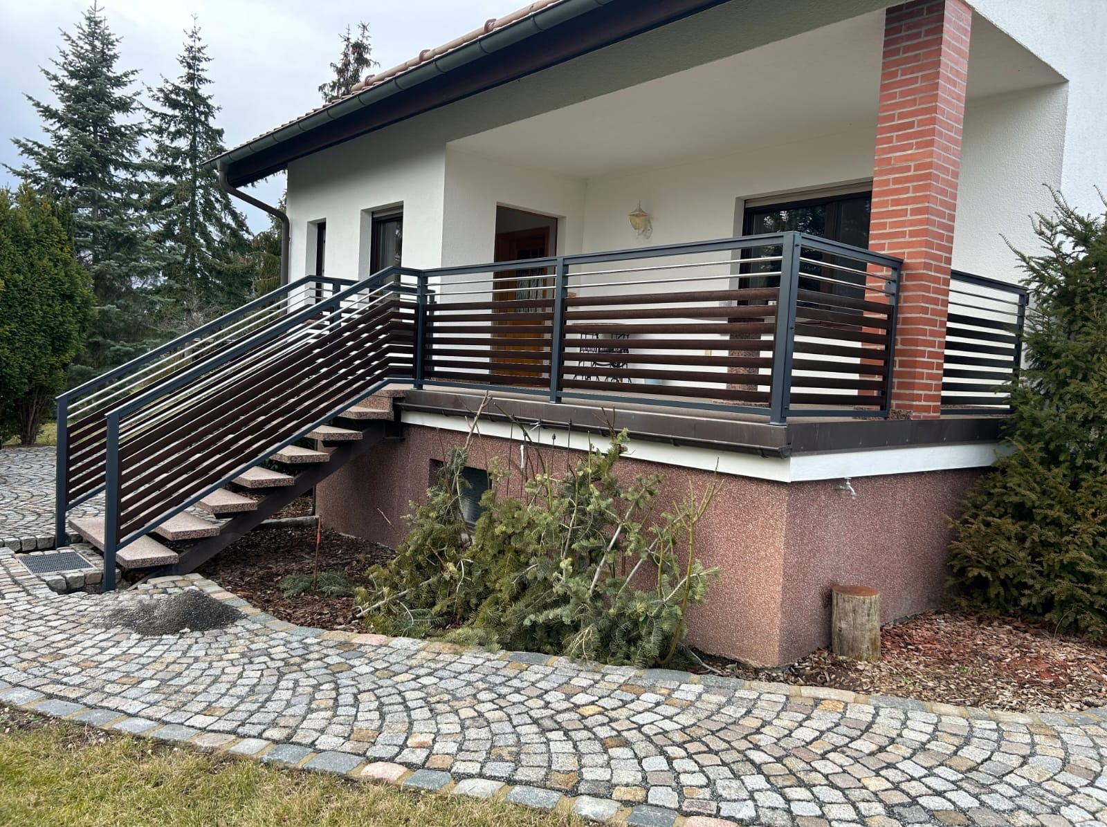 Nowoczesna balustrada balkonowa z poziomymi panelami i schodami, widok z brukowanego chodnika. Ciemna rama kontrastuje z jasną elewacją domu.