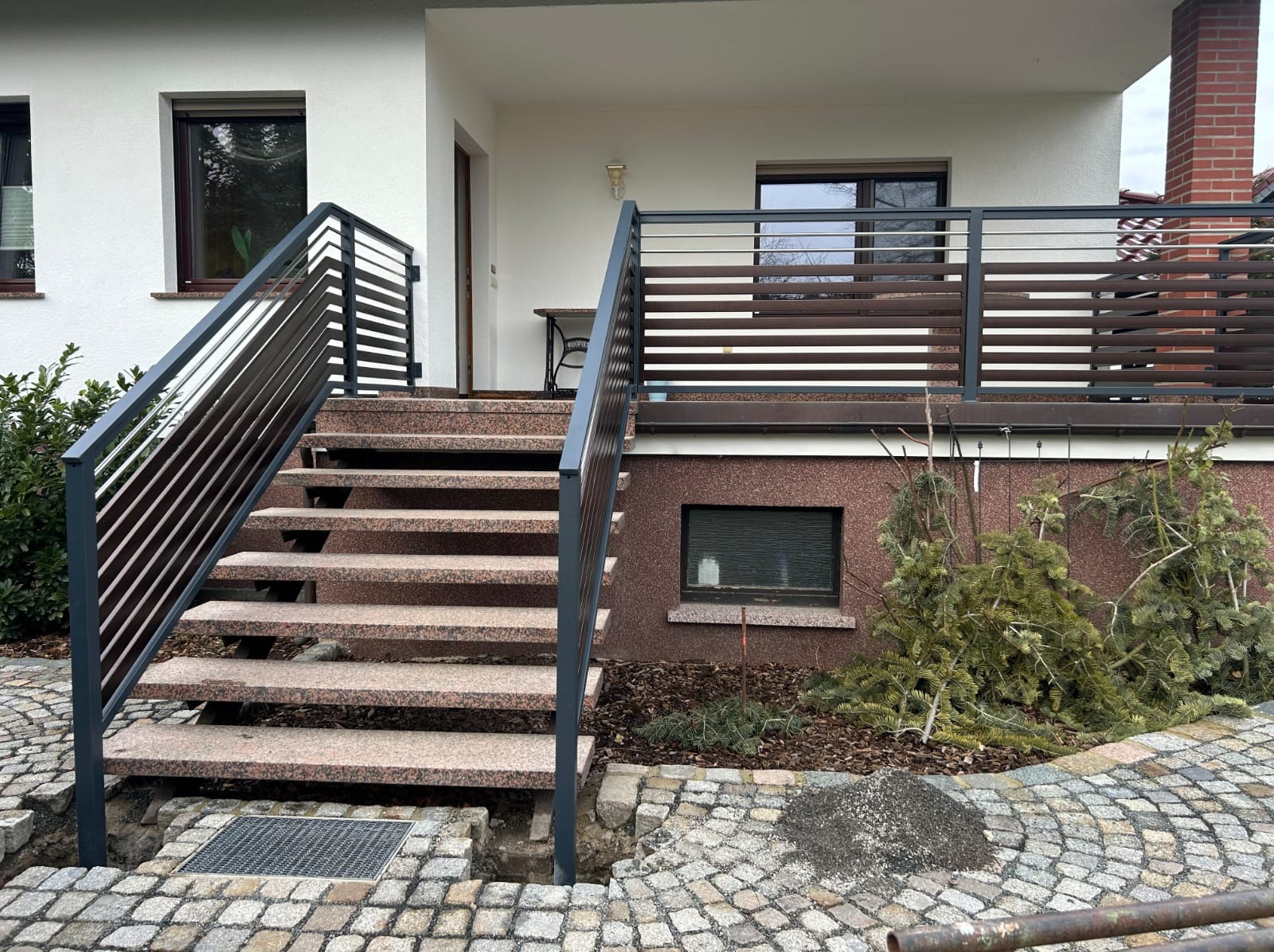 Schody wejściowe z różowego granitu z metalową balustradą w odcieniach szarości, prowadzące do balkonu z drewnianą barierką. Brukowana nawierzchnia.