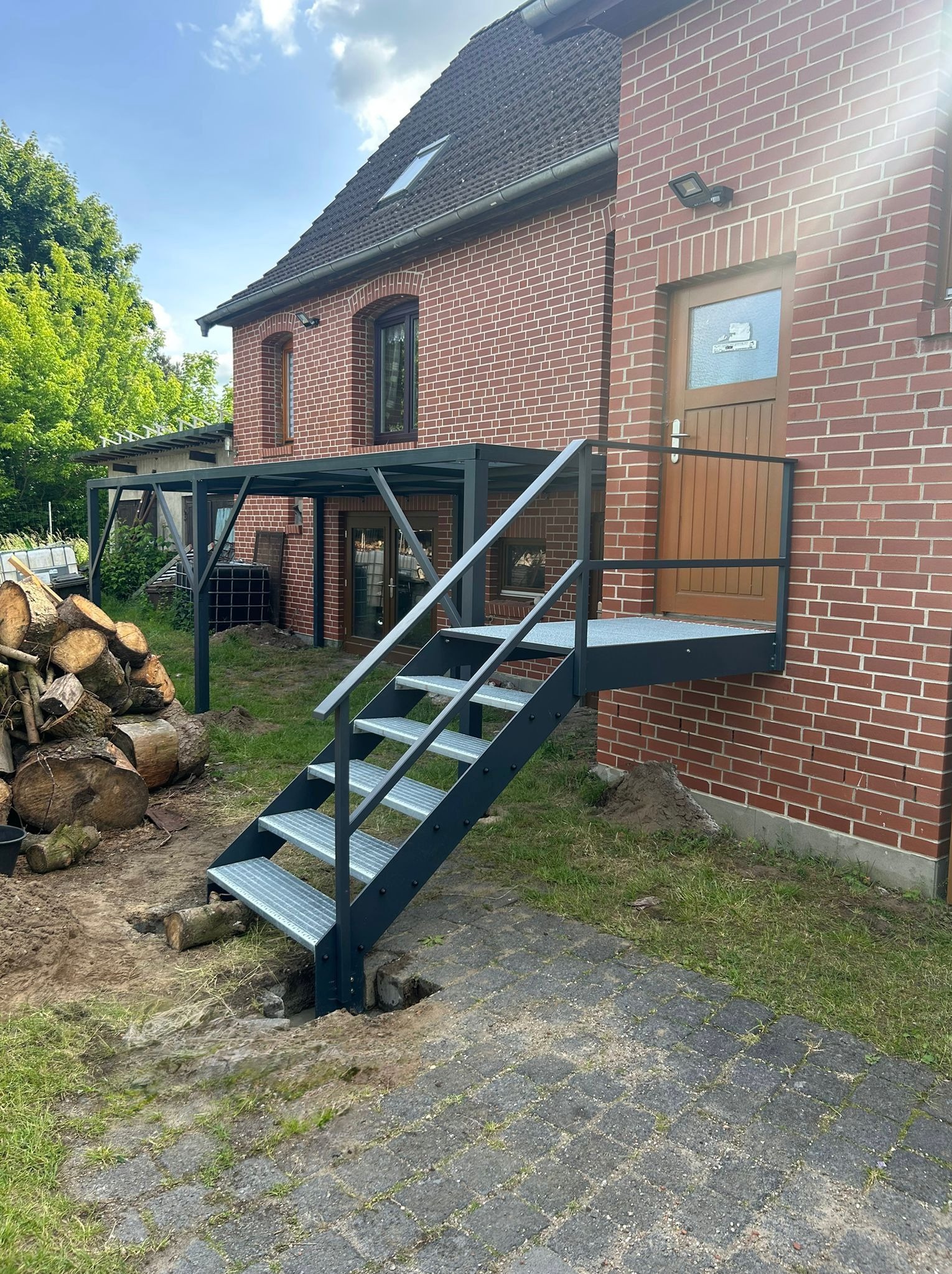 Zewnętrzne, metalowe schody z podestem prowadzą do drzwi w ceglanym budynku. W tle widoczna konstrukcja zadaszenia i ułożone drewno. Słoneczny dzień.