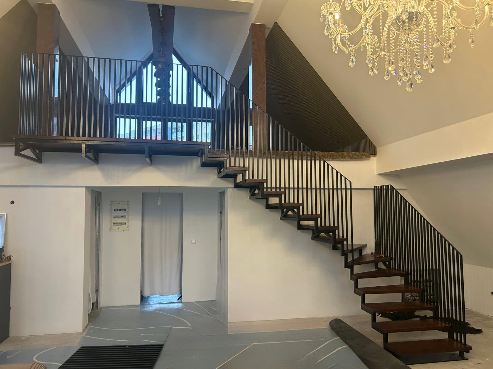 Nowoczesne schody z drewnianymi stopniami i czarną, metalową balustradą w przestronnym, wysokim wnętrzu z żyrandolem i antresolą.