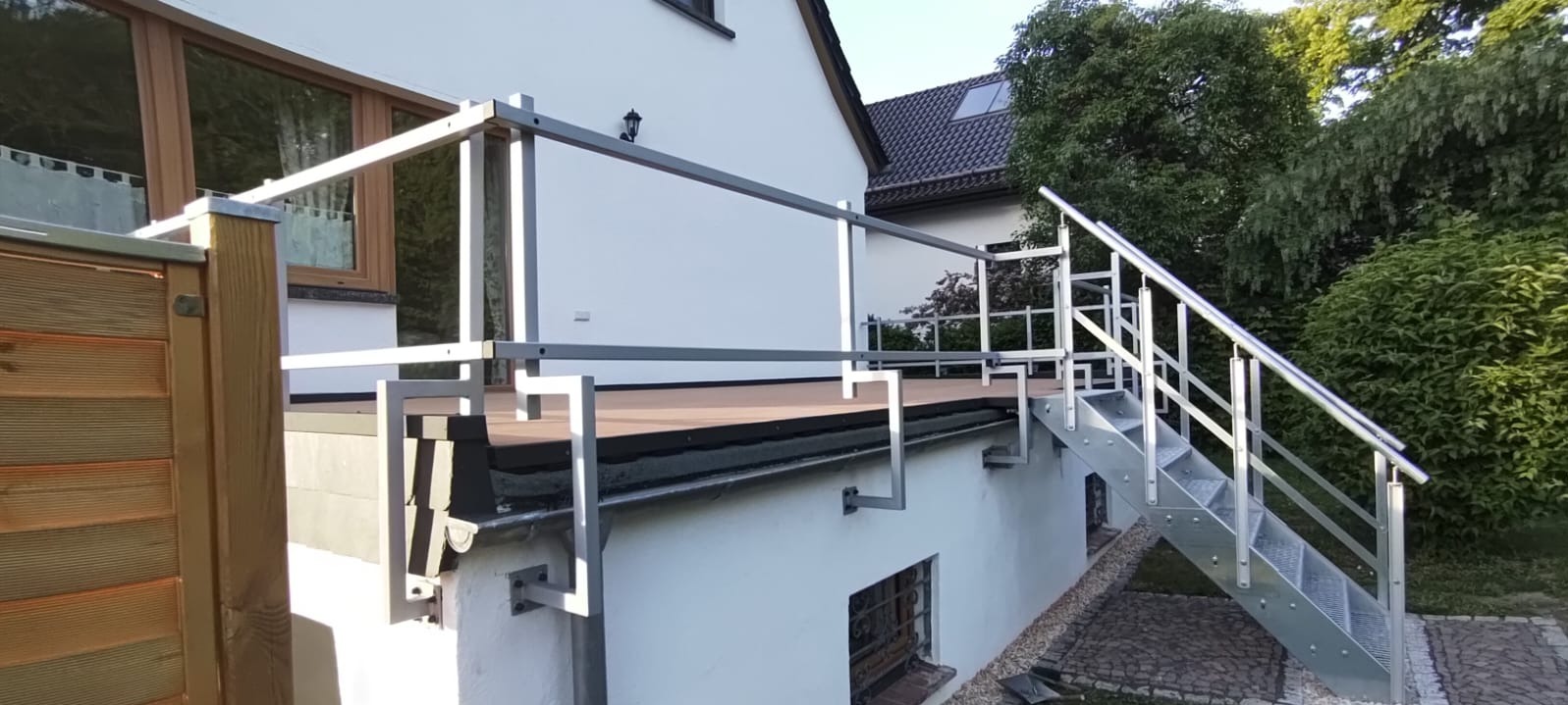 Metalowa balustrada z schodami na tarasie, widok z dołu, jasne kolory, dom w tle, zieleń. Solidna konstrukcja, nowoczesny design.