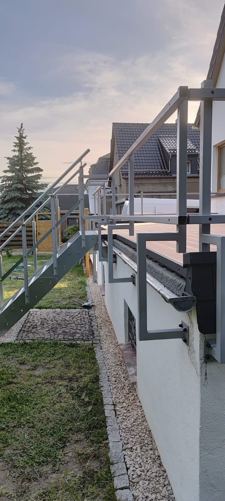 Szara metalowa balustrada z schodami na tarasie kompozytowym, widok z boku, w tle dom z ciemną dachówką i zielone drzewo iglaste.