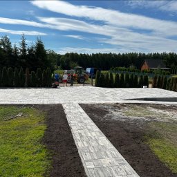 Usługi brukarskie -Łukasz Piwko - Jasnoszara kostka brukowa ułożona na podjeździe, widoczni pracownicy i maszyny budowlane na tle zieleni i błękitnego nieba. Widoczny szpadel na trawie.