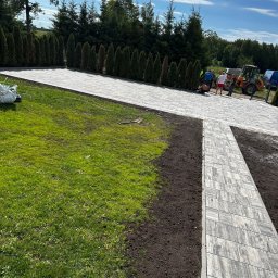 Usługi brukarskie -Łukasz Piwko - Kaskadowy układ szarej kostki brukowej na posesji z zielonym trawnikiem i rzędem tuj. W tle ekipa budowlana i sprzęt. Częściowo wykonana praca.