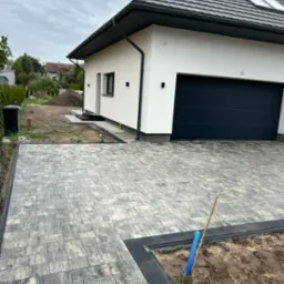 Ułożona kostka brukowa w szaro-beżowych odcieniach przed nowoczesnym domem z garażem. Czarna obwódka kostki, elementy drenażu i przygotowany teren pod nasadzenia.