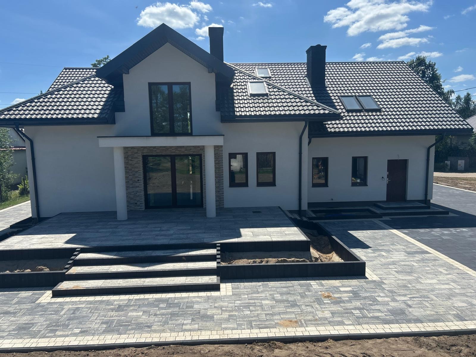 Nowoczesny dom jednorodzinny z szarym brukiem na podjeździe i tarasie. Widoczne schody z kostki i rabaty ziemne. Błękitne niebo w tle.
