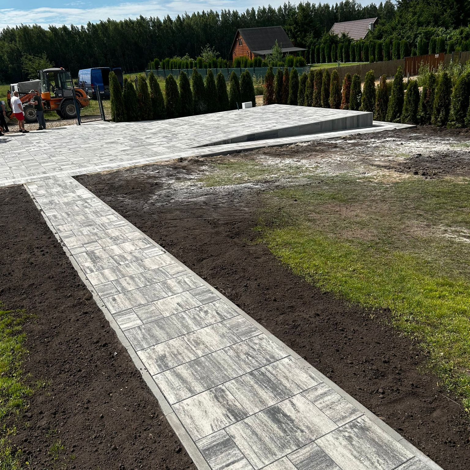 Wykończony chodnik z kostki brukowej w ogrodzie, z widocznym podjazdem i ekipą przy traktorze. Kostka w odcieniach szarości, ułożona w regularny wzór. Tło stanowią drzewa i budynki.