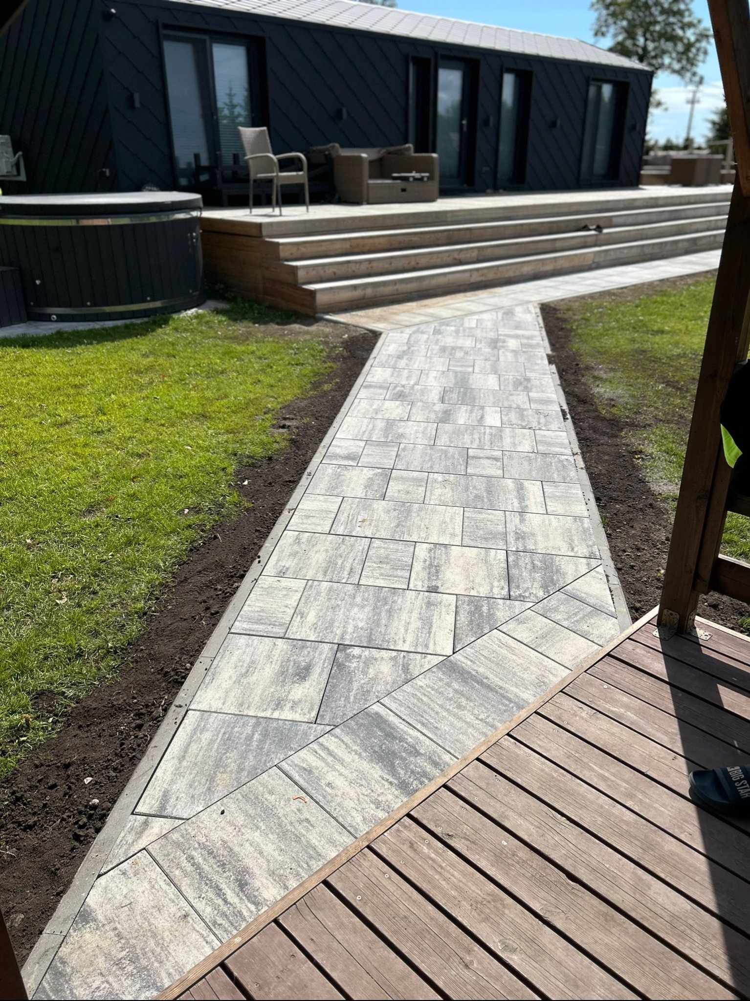 Ścieżka z jasnoszarej kostki brukowej prowadząca do nowoczesnego domu z czarną elewacją. Widoczny trawnik i fragment drewnianego tarasu.