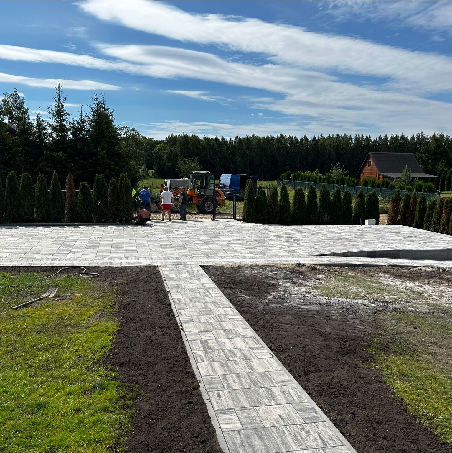 Jasnoszara kostka brukowa ułożona na podjeździe, widoczni pracownicy i maszyny budowlane na tle zieleni i błękitnego nieba. Widoczny szpadel na trawie.