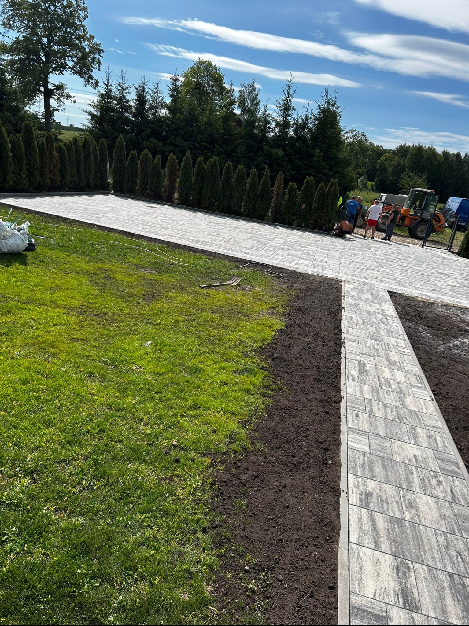 Kaskadowy układ szarej kostki brukowej na posesji z zielonym trawnikiem i rzędem tuj. W tle ekipa budowlana i sprzęt. Częściowo wykonana praca.