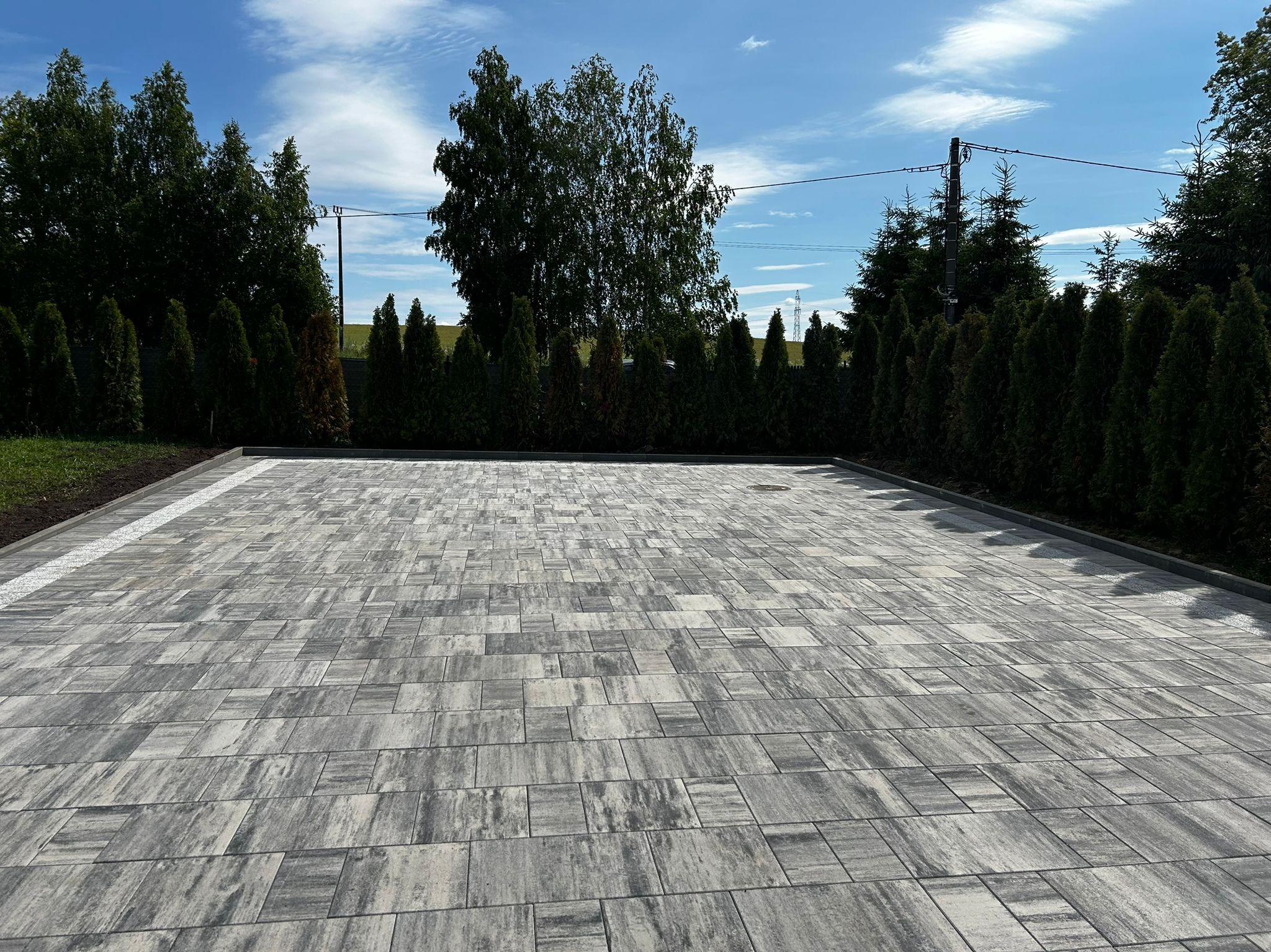 Ułożona szara kostka brukowa na posesji, obrzeża wykończone drobnym kamieniem. W tle rząd tuj i drzewa liściaste pod błękitnym niebem.
