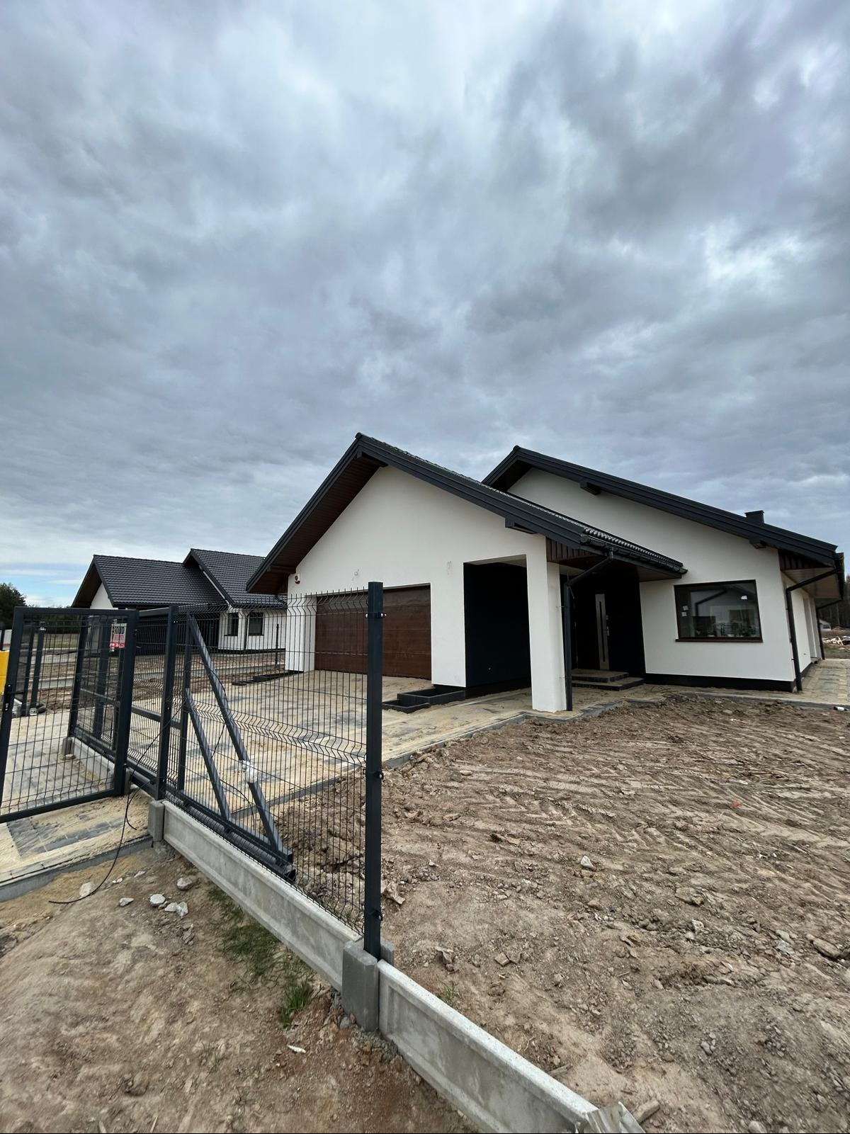 Nowoczesny dom jednorodzinny z grafitową bramą wjazdową i ogrodzeniem panelowym na niezagospodarowanej działce. Widok na elewację i podjazd z kostki.