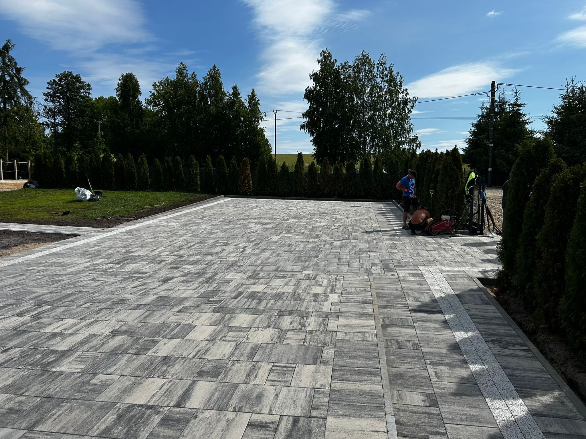 Ukończona nawierzchnia z szarej kostki brukowej, obrzeża z jasnej kostki. W tle zieleń drzew i krzewów, pracownicy przy maszynach. Widok na posesję po pracach.