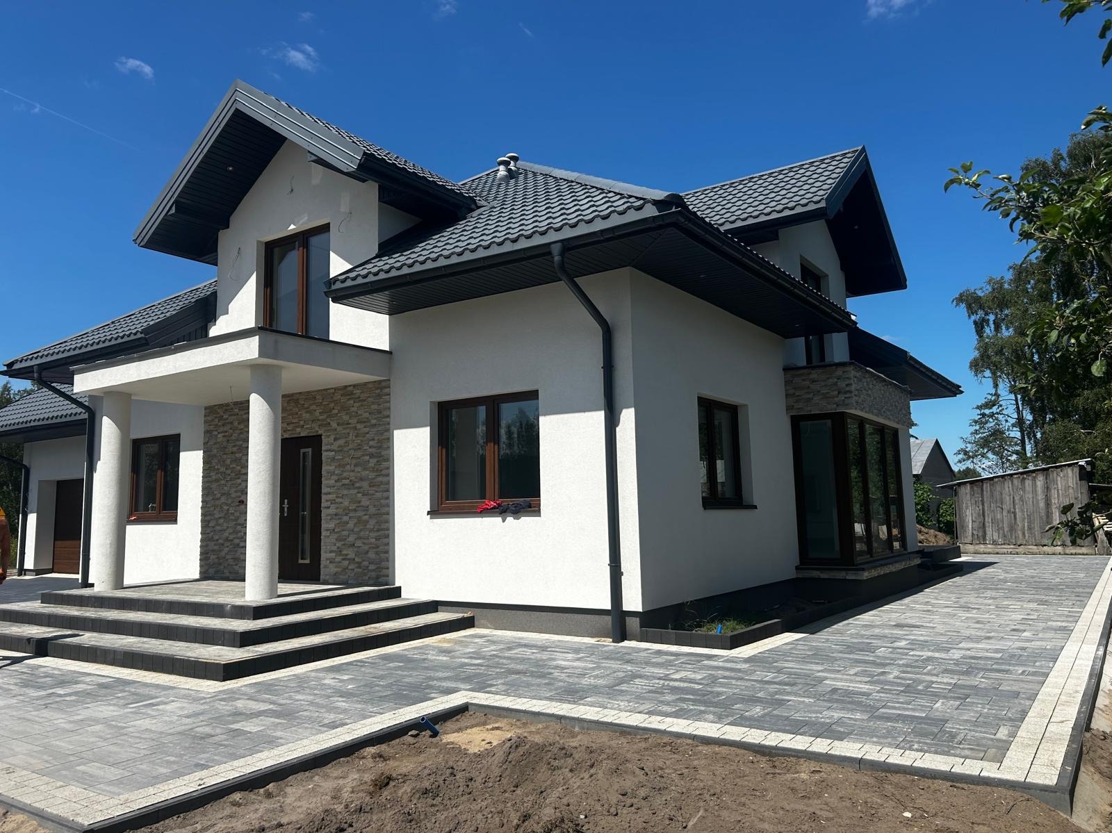 Elegancki dom jednorodzinny z szarą kostką brukową na podjeździe i schodach wejściowych, jasna elewacja, ciemna dachówka, błękitne niebo w tle.