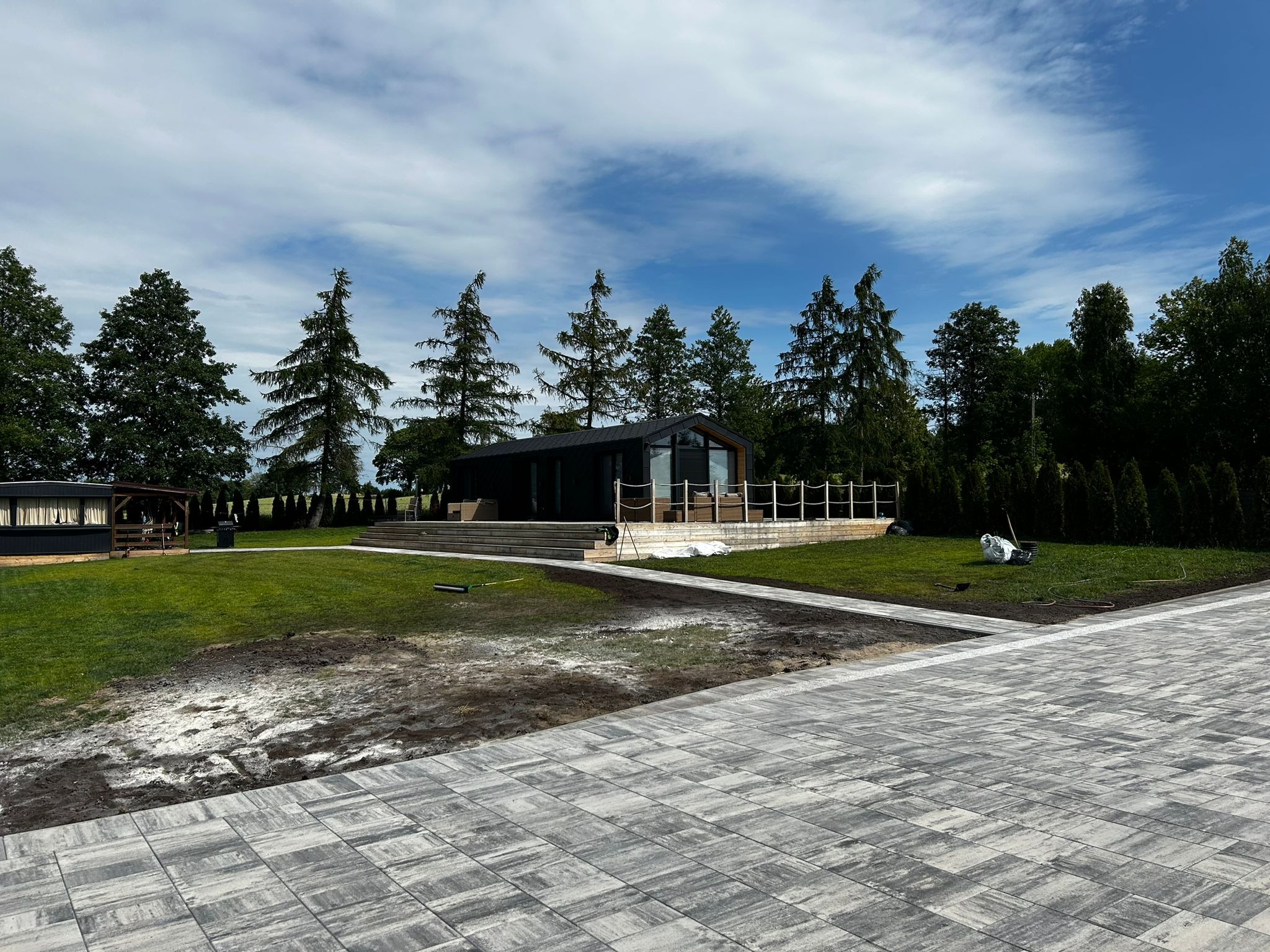 Nowoczesny dom z ciemną elewacją i tarasem, otoczony zielonym trawnikiem oraz ścieżką z szarej kostki brukowej. Częściowo przygotowany teren pod nasadzenia.