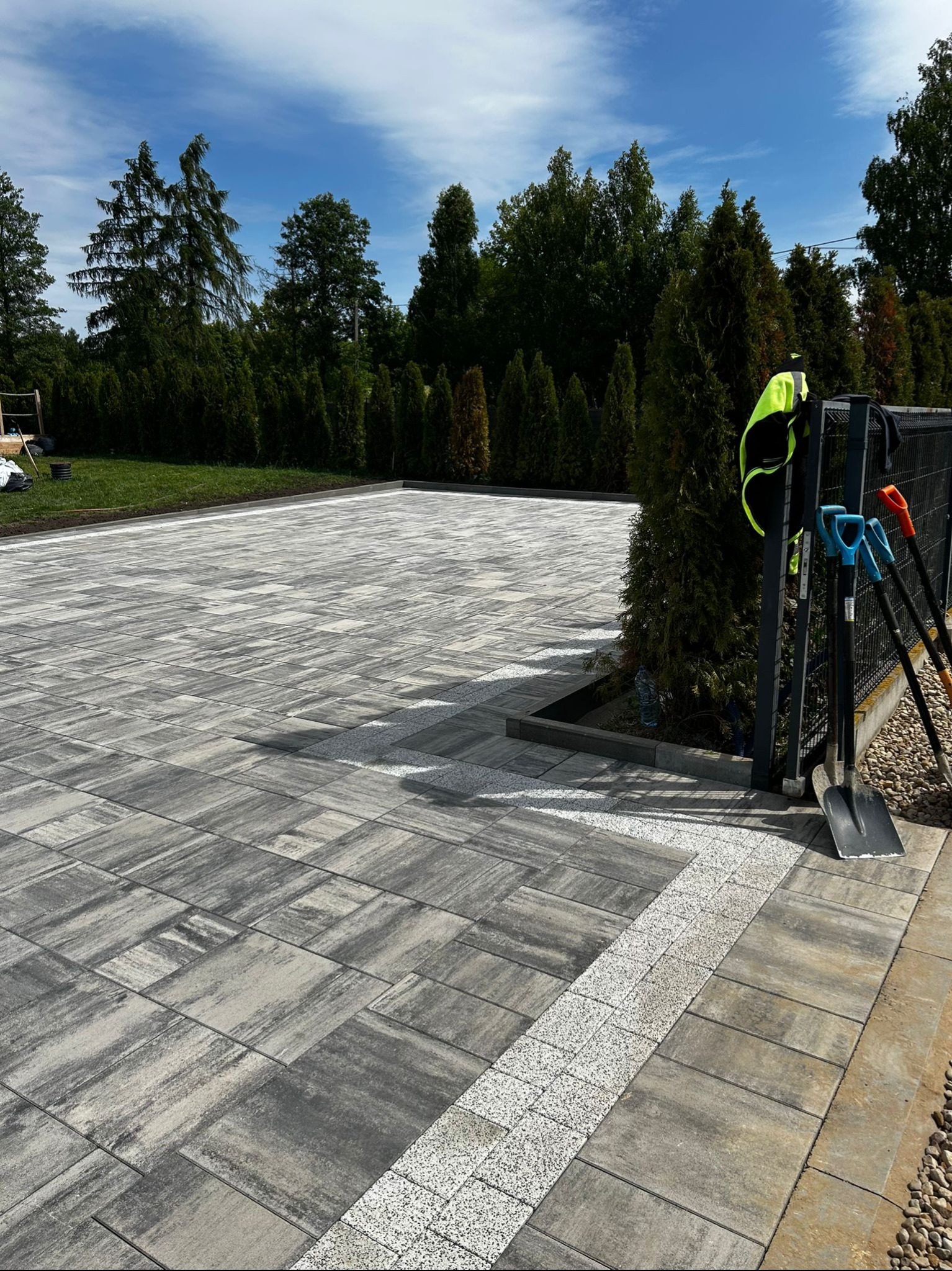 Ukończona nawierzchnia z szarej kostki brukowej, obrzeże z drobnego kruszywa. Przy ogrodzeniu narzędzia: łopaty i kamizelka odblaskowa. Zieleń w tle.