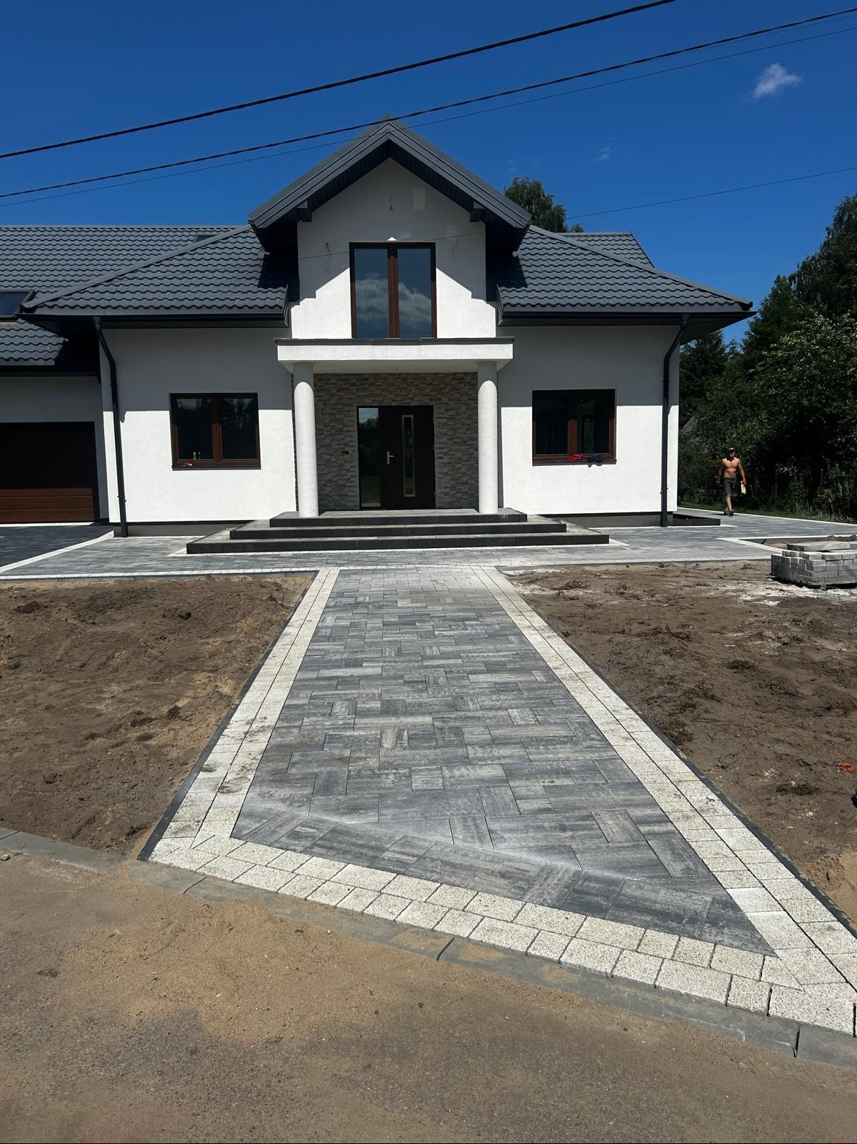 Nowoczesny podjazd z kostki brukowej w odcieniach szarości prowadzący do domu z białej elewacji i ciemnym dachem. Widoczne obrzeża i przygotowany teren pod trawnik.