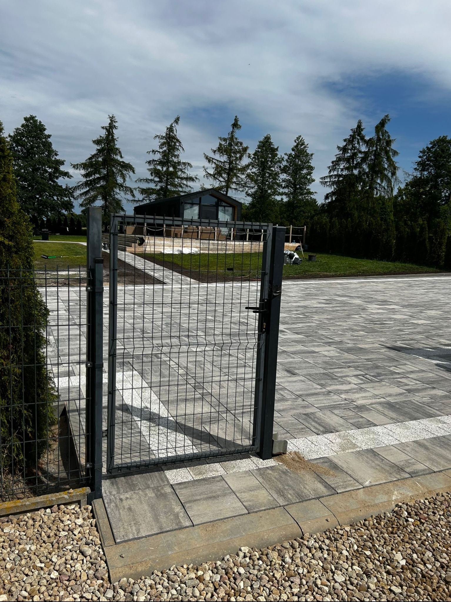 Nowoczesna posesja: grafitowa siatka ogrodzeniowa z furtką, jasnoszara kostka brukowa z białymi akcentami, dom w tle otoczony zielenią i drzewami.