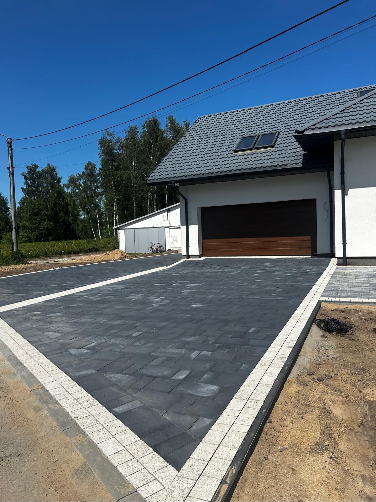 Nowoczesny podjazd z ciemnej kostki brukowej obramowanej jaśniejszą kostką, prowadzący do garażu domu jednorodzinnego w słoneczny dzień. Widoczne detale wykończenia.