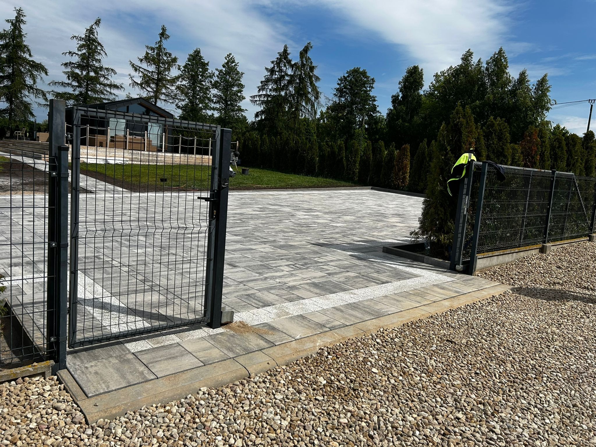Nowoczesny podjazd z kostki brukowej w odcieniach szarości, obrzeża z jasnych kostek, otoczony żwirem i ogrodzeniem z siatki. W tle widoczny budynek.