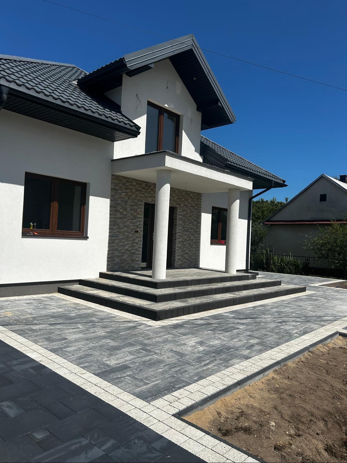 Nowoczesny dom z eleganckim wejściem wyłożonym szarą kostką brukową z jasną obwódką. Schody prowadzą do wejścia z kolumnami. Czyste, błękitne niebo w tle.
