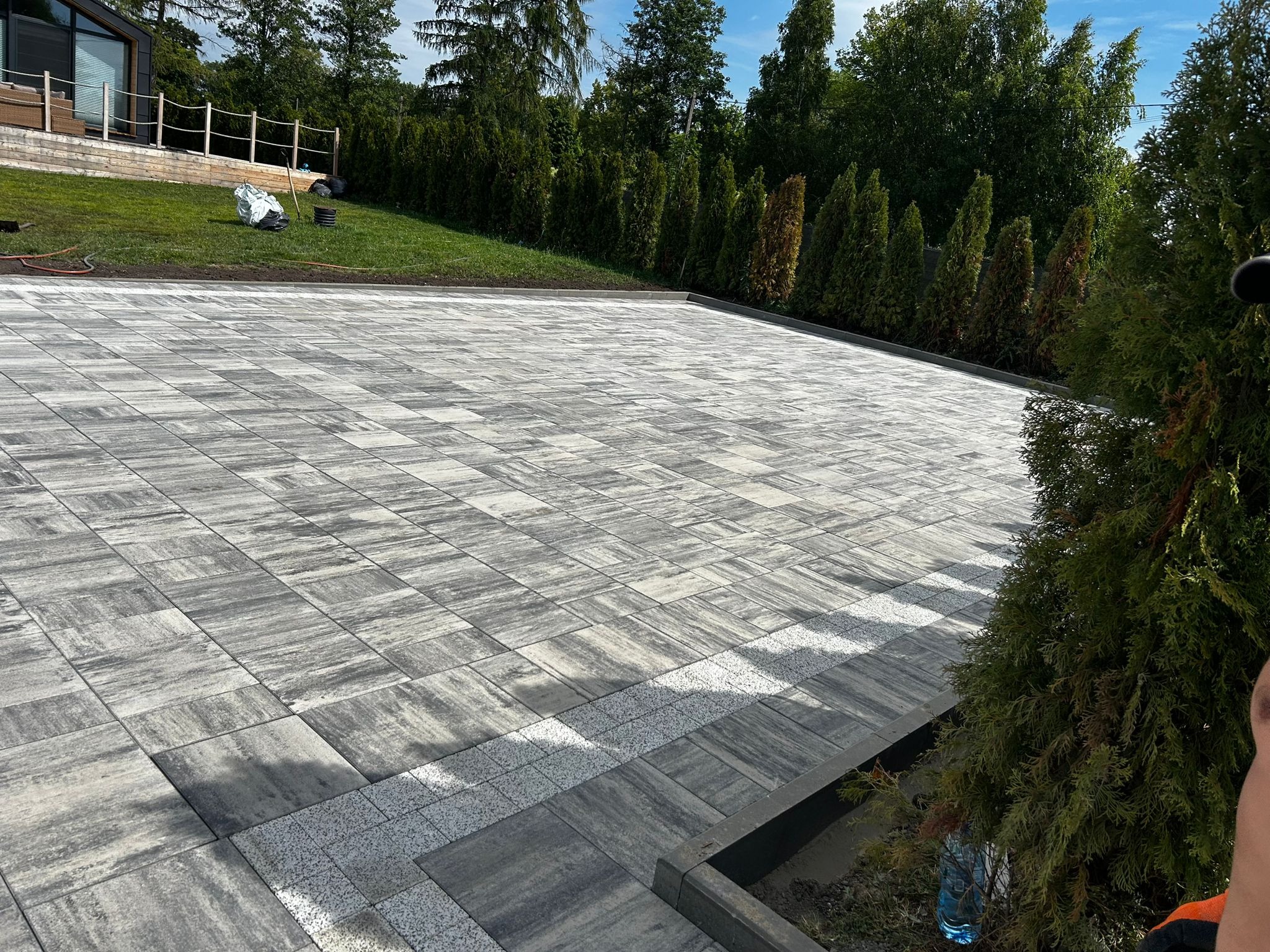Ułożona kostka brukowa w odcieniach szarości, z dekoracyjnym pasem z drobnej kostki. W tle widoczny dom z drewnianą elewacją i zielony trawnik.