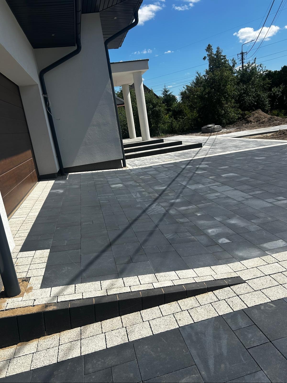 Nowoczesna, grafitowa kostka brukowa z jasną obwódką przed domem z kolumnami. Widoczne schody wejściowe i brama garażowa. Słoneczny dzień.