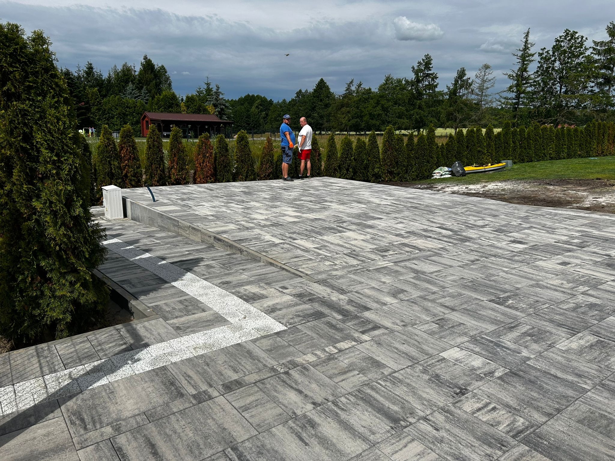 Wykończony taras z kostki brukowej w odcieniach szarości, z dekoracyjnymi białymi elementami. W tle drzewa i dwóch mężczyzn rozmawiających na tle zieleni.