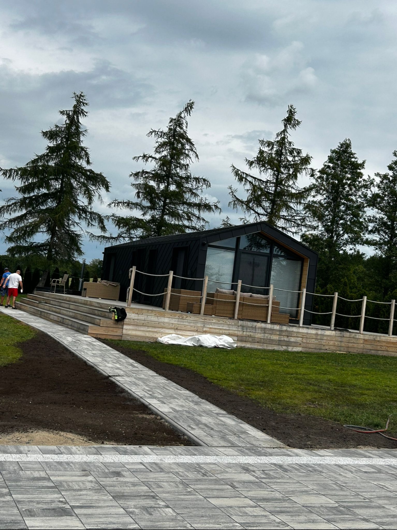 Ułożona szara kostka brukowa prowadząca do nowoczesnego domu z drewnianym tarasem. Widoczne drzewa iglaste i fragment trawnika.