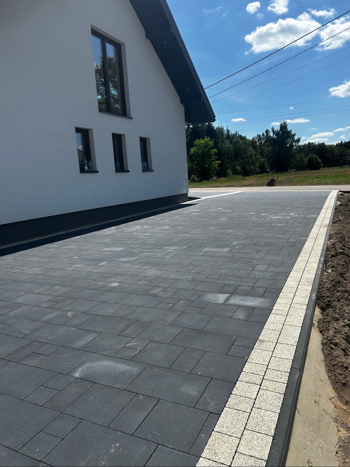 Ułożona, grafitowa kostka brukowa z jasnym obrzeżem na podjeździe przed nowoczesnym domem. Widoczne ślady po deszczu na kostce i świeżo obsypana ziemia po bokach.