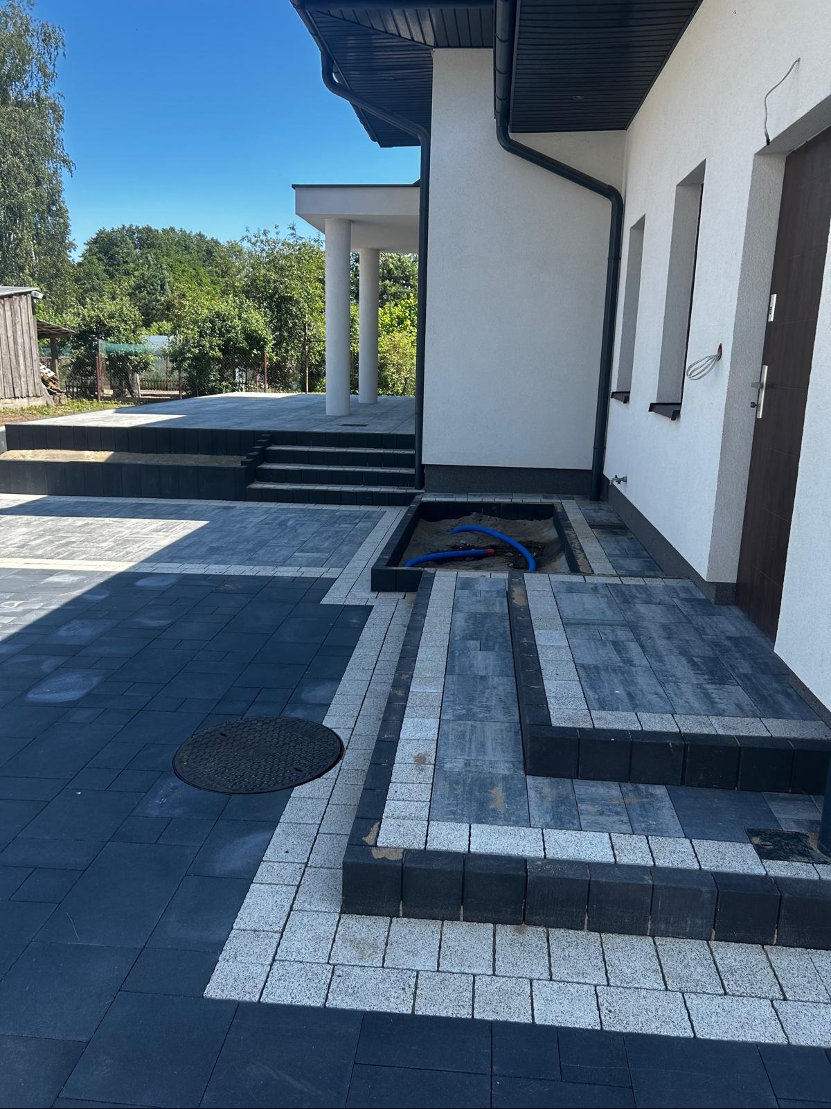 Nowoczesna posesja z czarno-szarą kostką brukową, schodami i studzienką. W tle widoczny budynek z kolumnami i fragment zieleni. Częściowo widoczne niebieskie rury.