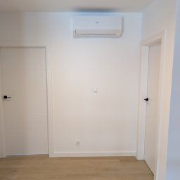 Simply Green Dorota Cierniewska - Biała klimatyzacja Haier zamontowana na ścianie w nowoczesnym wnętrzu z dwiema białymi drzwiami i drewnianą podłogą. Minimalistyczny design.