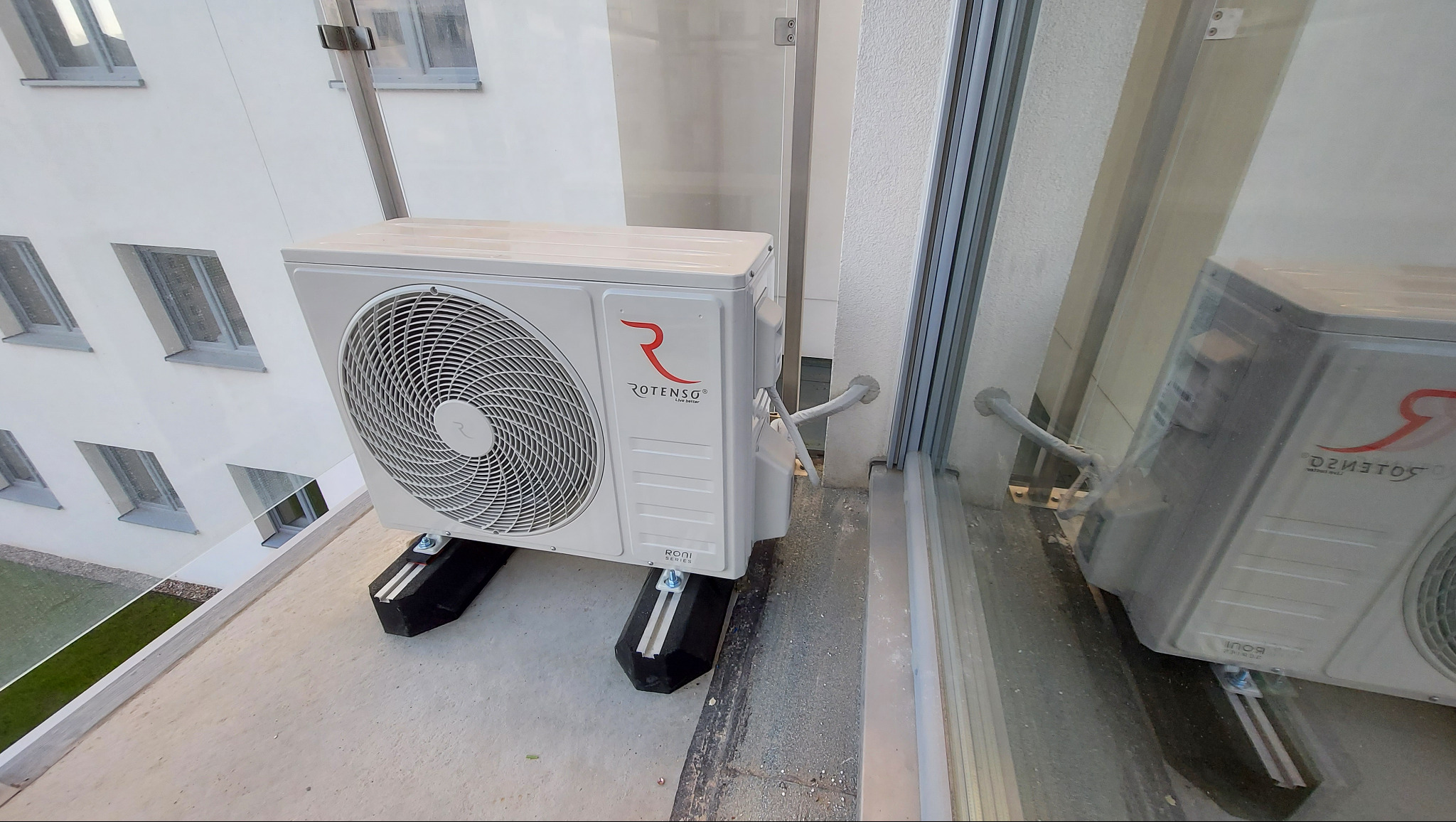 Biała jednostka zewnętrzna klimatyzacji Rotenso zamontowana na wspornikach na balkonie, z widokiem na okna budynku w tle i odbiciem w szybie.