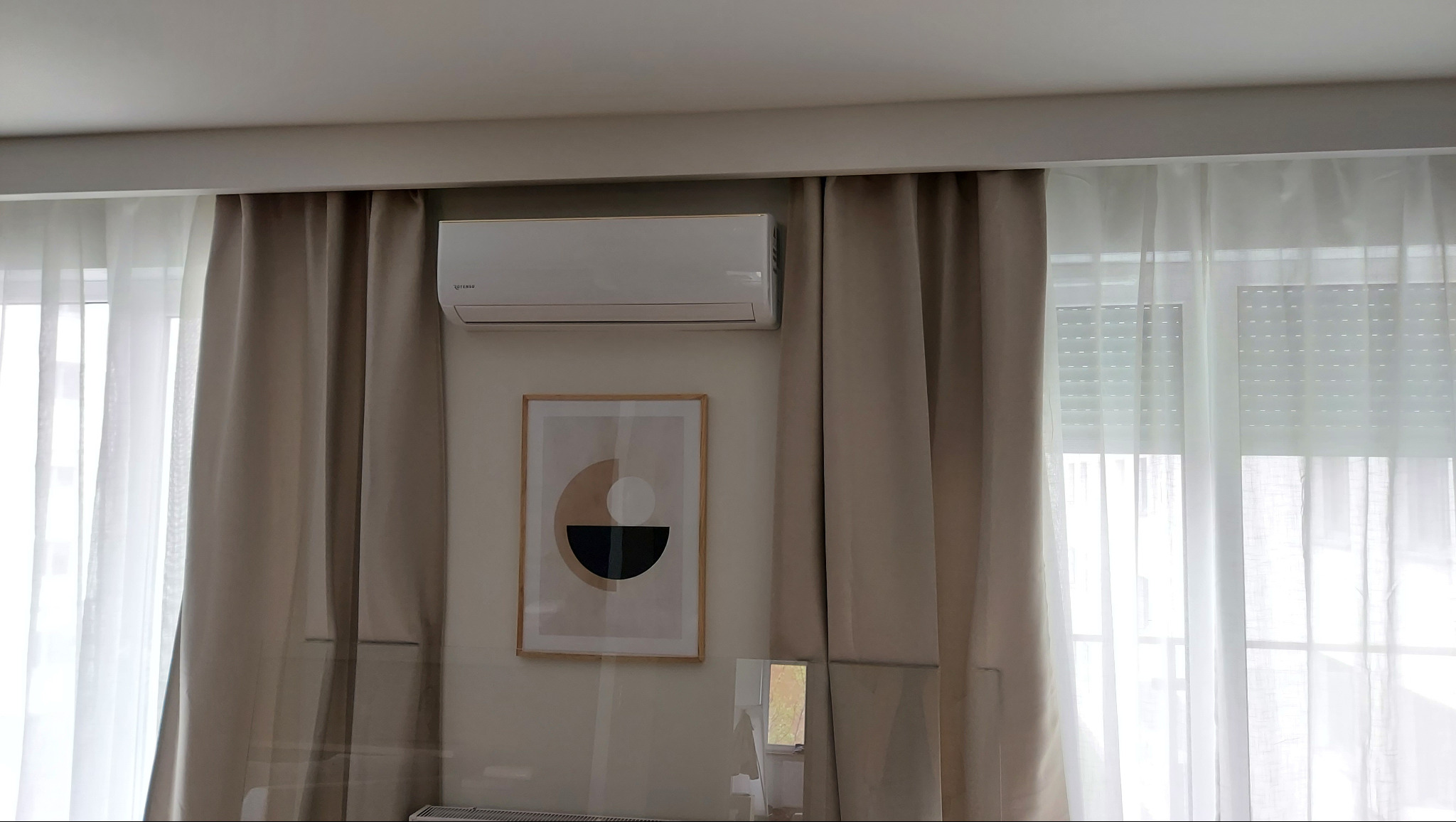 Biała klimatyzacja na ścianie nad obrazem w ramie, obok zasłoniętego okna. Neutralna kolorystyka wnętrza, minimalistyczny design.