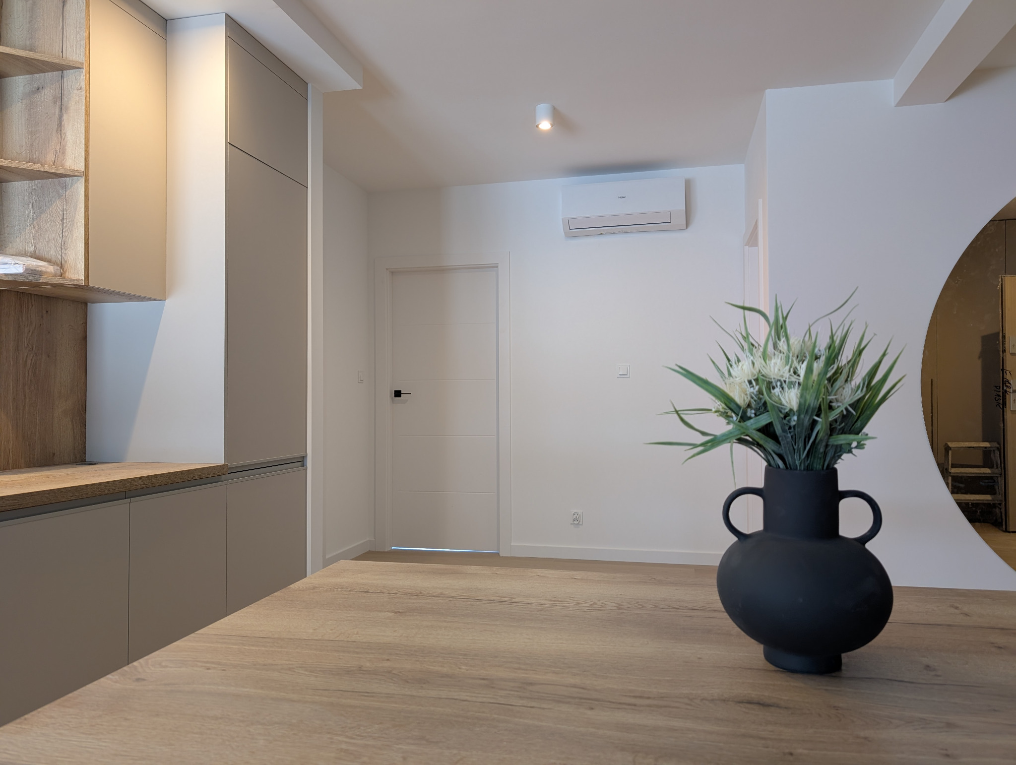Wnętrze domu z zamontowaną klimatyzacją Haier na ścianie nad drzwiami. Minimalistyczny design, jasne kolory, drewniany blat i dekoracyjny wazon.